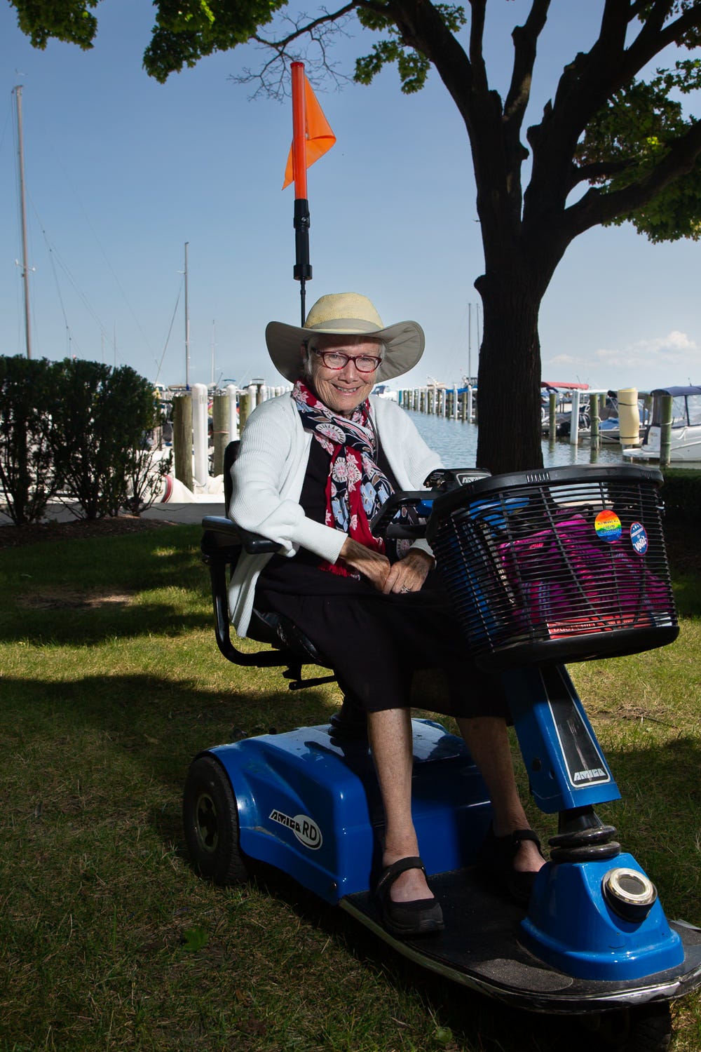 Patricia Lay-Dorsey smiles while sitting on a blue mobility scooter beneath a large tree at a marina. She wears a wide-brimmed hat, glasses, a patterned scarf, and a white cardigan. Boats and docks are visible behind her on a sunny day. Patricia Lay-Dorsey smiles while sitting on a blue mobility scooter beneath a large tree at a marina. She wears a wide-brimmed hat, glasses, a patterned scarf, and a white cardigan. Boats and docks are visible behind her on a sunny day.