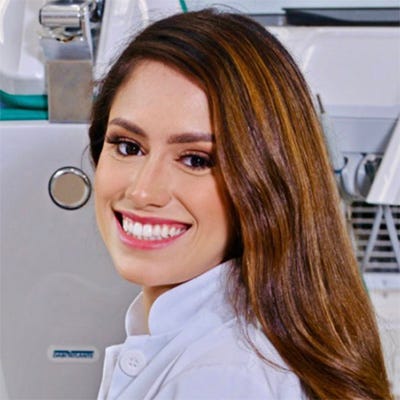 A foto mostra uma mulher jovem, de jaleco branco, sorrindo em um ambiente de laboratório, com equipamentos científicos ao fundo.