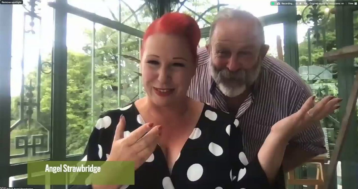 Smiling Angel Strawbridge with red hair and polka dot dress gestures while her husband Dick Strawbridge, a bearded man with a striped shirt stands behind her.  They are in a garden room surrounded by greenery.