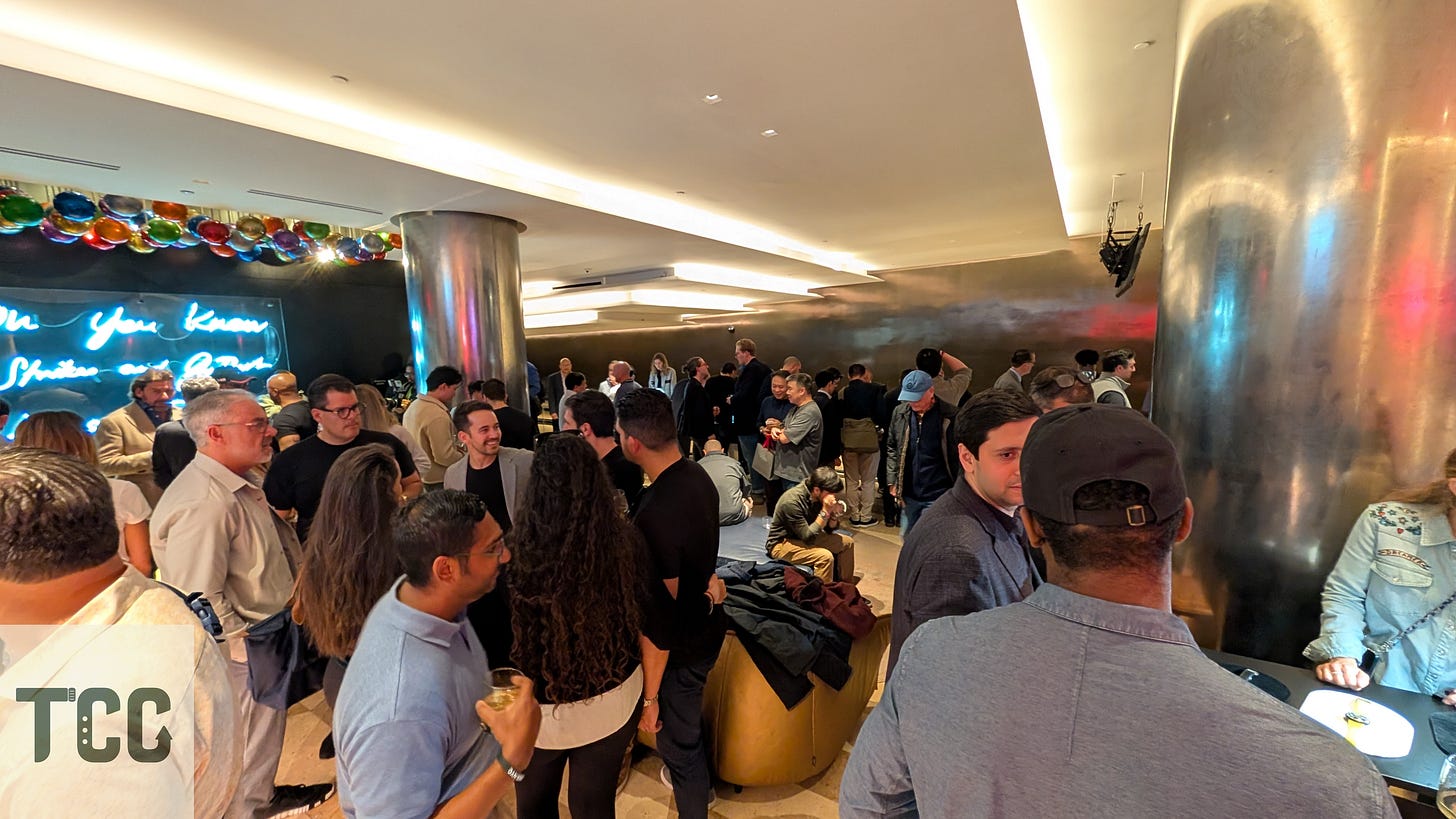 Another angle of the lively crowd at The Indies, showing people engaged in animated conversations and watch demonstrations under soft, moody lighting. A neon sign and metallic walls add atmosphere to the venue.
