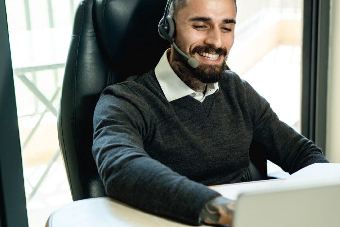 A man sitting at an office desk in business casual, smiling and having a headset on while looking at a laptop. A man sitting at an office desk in business casual, smiling and having a headset on while looking at a laptop.