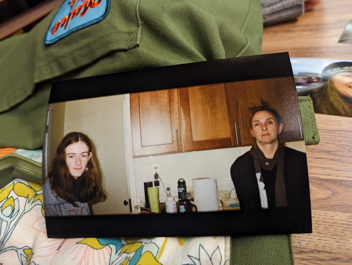 A film photo of a teenager and her mother (the author of this essay) in a cluttered kitchen (why are there so many water bottles?). The photo is propped against a handmade bag, on the table of a Chinese restaurant, where we were waiting for our order. A film photo of a teenager and her mother (the author of this essay) in a cluttered kitchen (why are there so many water bottles?). The photo is propped against a handmade bag, on the table of a Chinese restaurant, where we were waiting for our order.