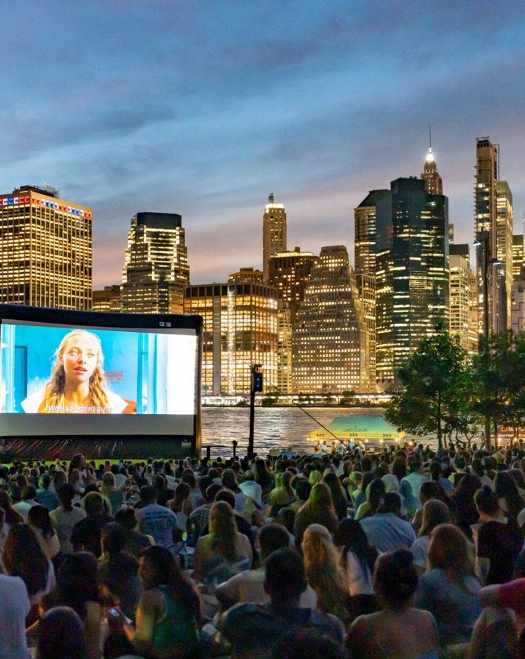 This may contain: a large group of people sitting in front of a city skyline watching a movie on an outdoor projector screen