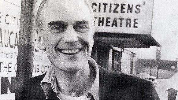 A black and white shot of a man outsizxe the Citizens' Theatre in Glasgow. He is clean shaven with short hair and has a wide smile. He wears a dark blazer, a shirt unbuttoned at the neck and a light t-shirt, with a Citizens' Theatre badge on his lapel.