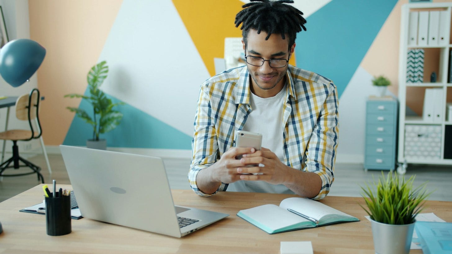 A man in glasses and a plaid shirt looks at his phone while sitting at a desk with an open laptop, a notebook and a small plant. A man in glasses and a plaid shirt looks at his phone while sitting at a desk with an open laptop, a notebook and a small plant.