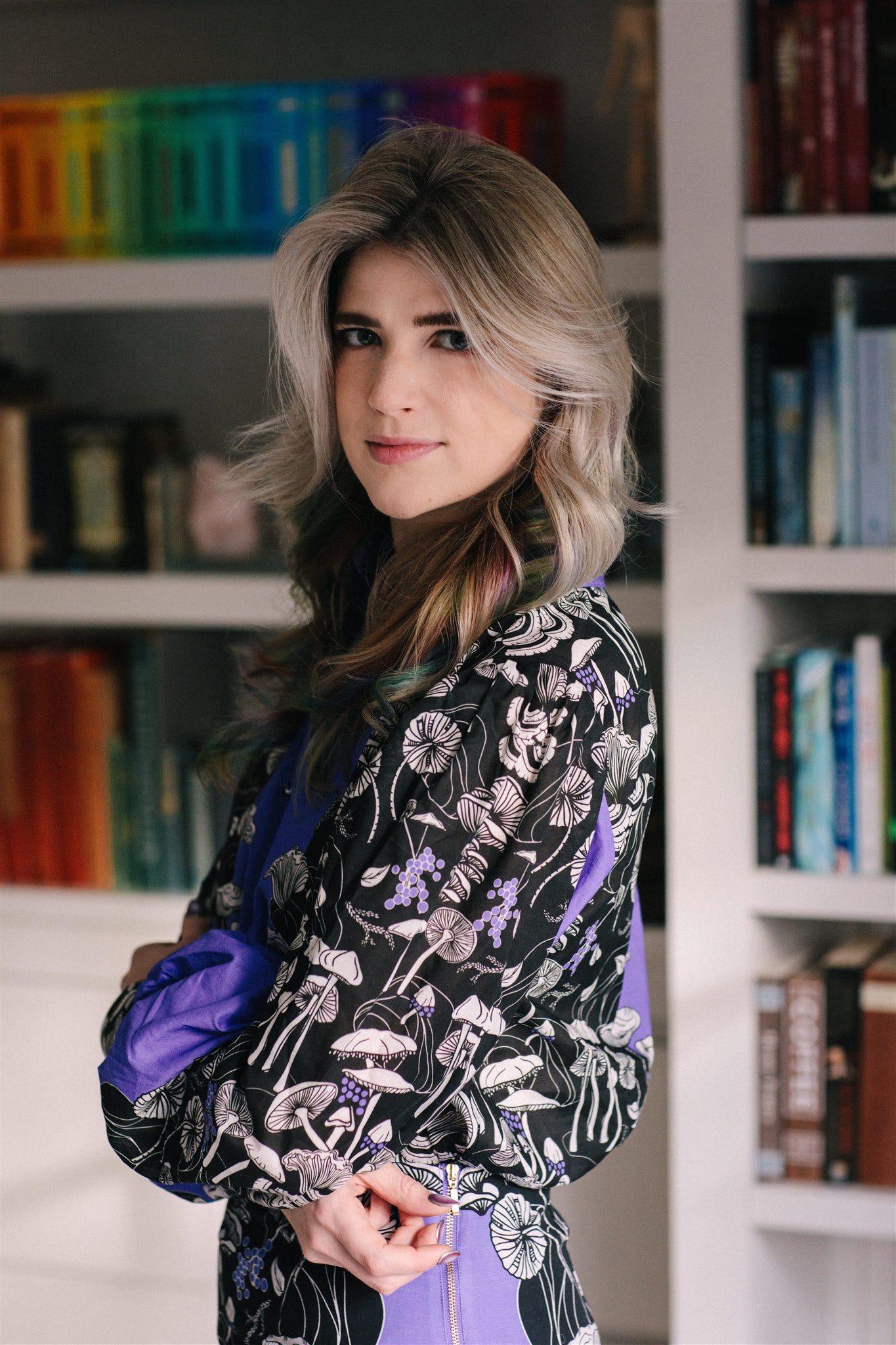 A photo of Taylor, a blonde and rainbow haired femme, in a purple ensemble featuring a pattern of mushrooms, in front of rainbow books, looking over their shoulder at the camera