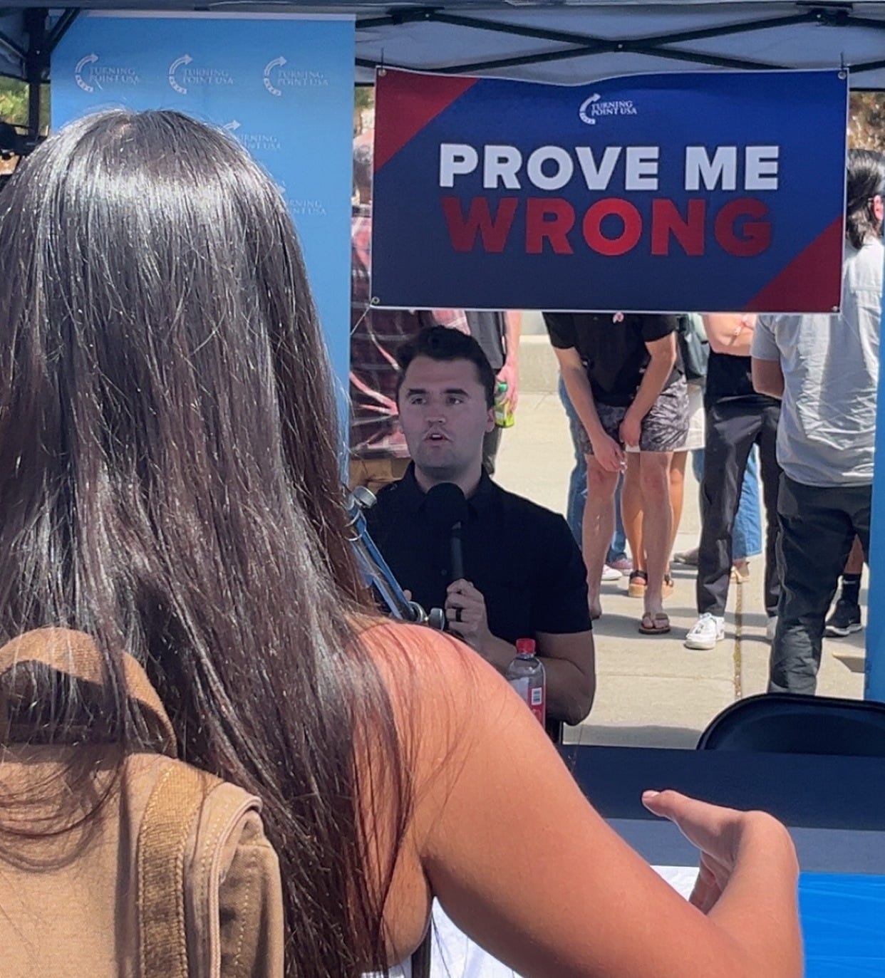 Kirk under a sign saying Prove Me Wrong; a student is walking towards him, long hair, presumably to debate him. he holds a microphone. Kirk under a sign saying Prove Me Wrong; a student is walking towards him, long hair, presumably to debate him. he holds a microphone.