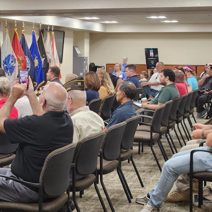 The image shows a formal indoor event, likely honoring veterans or related to military service. A man in a suit stands at a podium, speaking to an audience. Behind him are multiple flags, including the United States flag and the black-and-white POW/MIA flag. There is also a backdrop with the logo and text "FHP" and "Florida Highway Patrol." Audience members, some wearing veteran caps and uniforms, are seated, with one person in a blue shirt that reads "MSGT SOUTH." The general atmosphere is respectful and attentive.