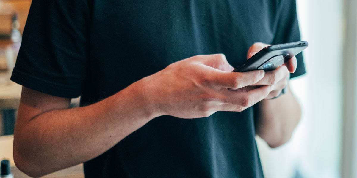 Foto de um homem branco com camiseta preta segurando um smartphone.