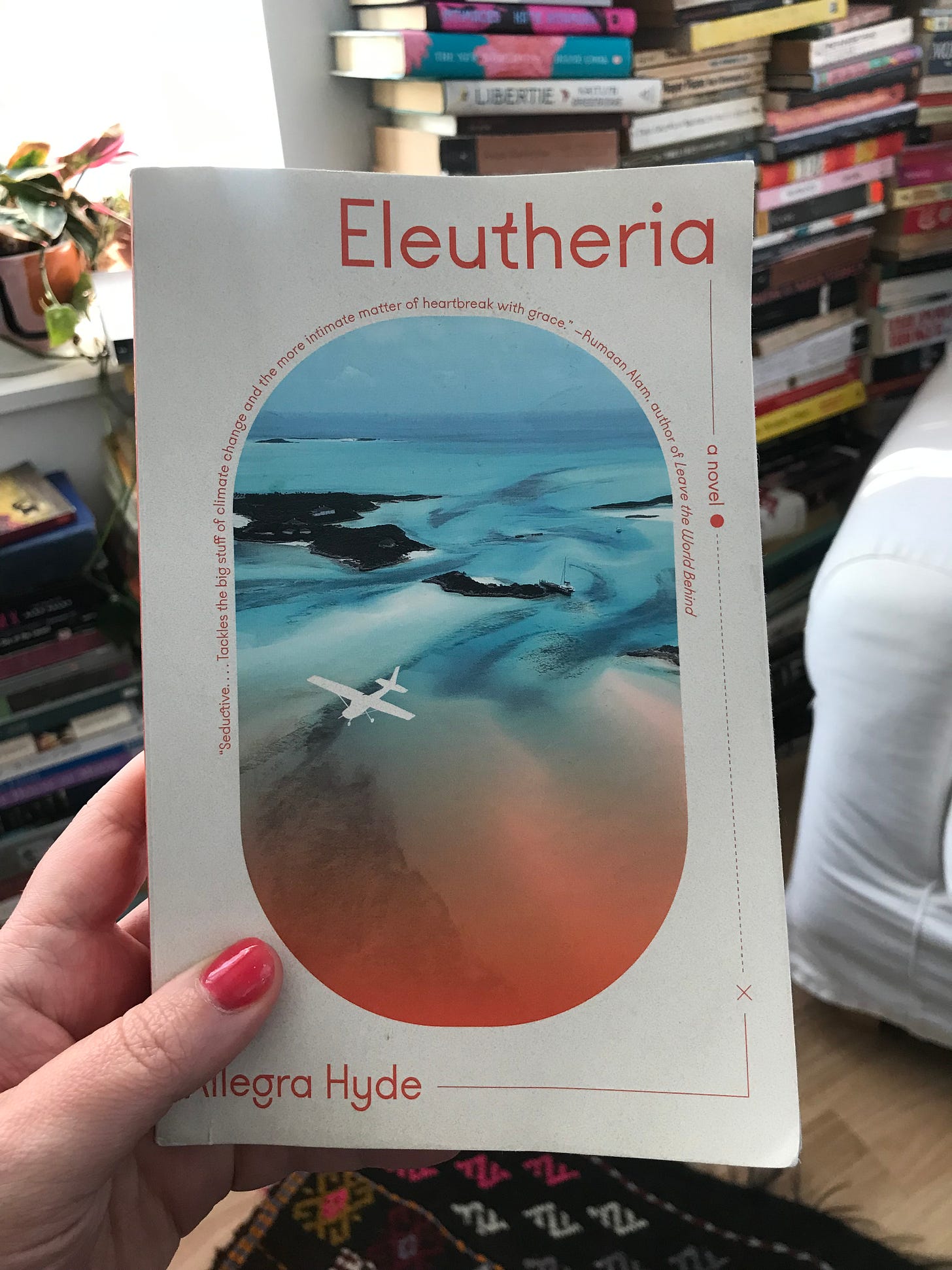 Hand holding a book called Eleutheria, which has a cover depicting a Caribbean island and a small plane. Behind the book are plants and stacks of other books.