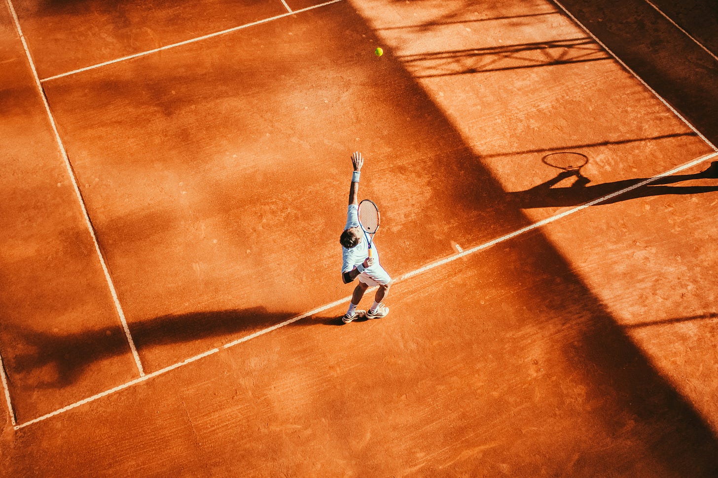 A tennis player wearing all white prepares to serve on a clay court.