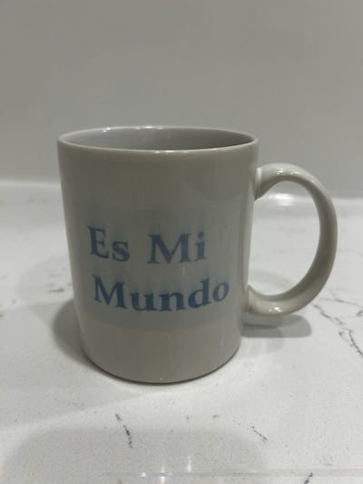 Photograph of a white mug with blue lettering. It reads, "Es Mi Mundo." Photograph of a white mug with blue lettering. It reads, "Es Mi Mundo."