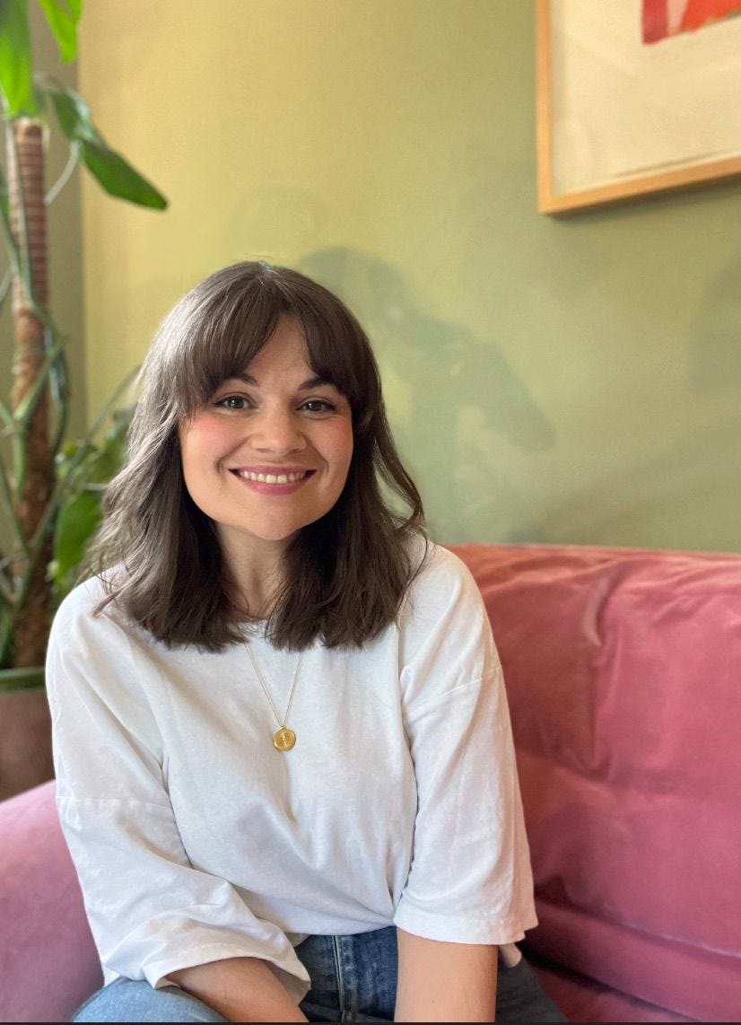 A picture of Lauren Farnsworth seated on a soft pink sofa. Her brown hair is in a long bob and she's wearing jeans and a white top. There is a houseplant and a picture frame in the background