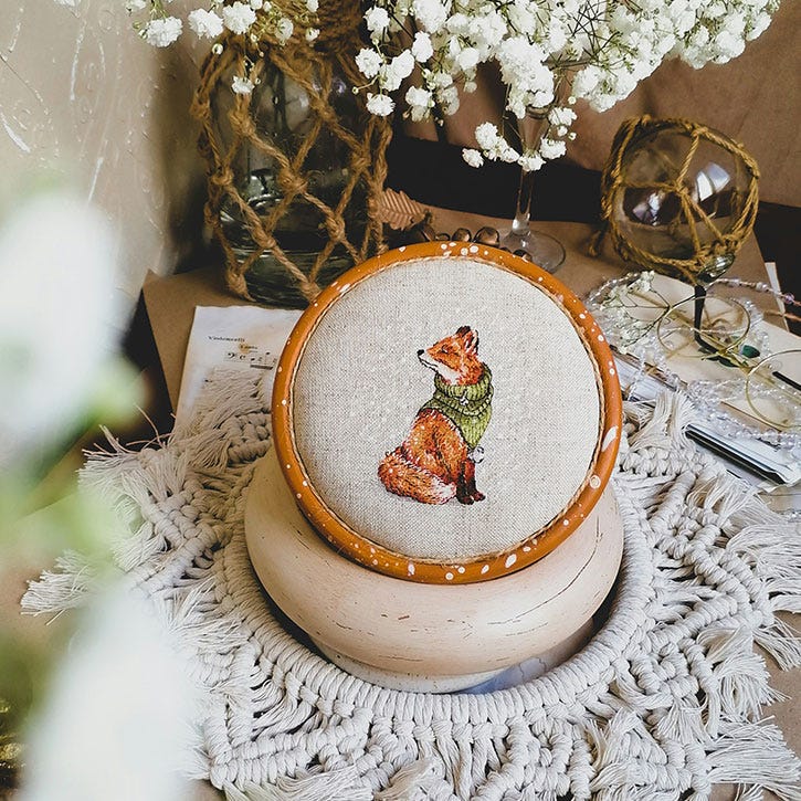 A cute little cross-stitch of a fox wearing a scarf on a table topped with a vase filled with white flowers A cute little cross-stitch of a fox wearing a scarf on a table topped with a vase filled with white flowers