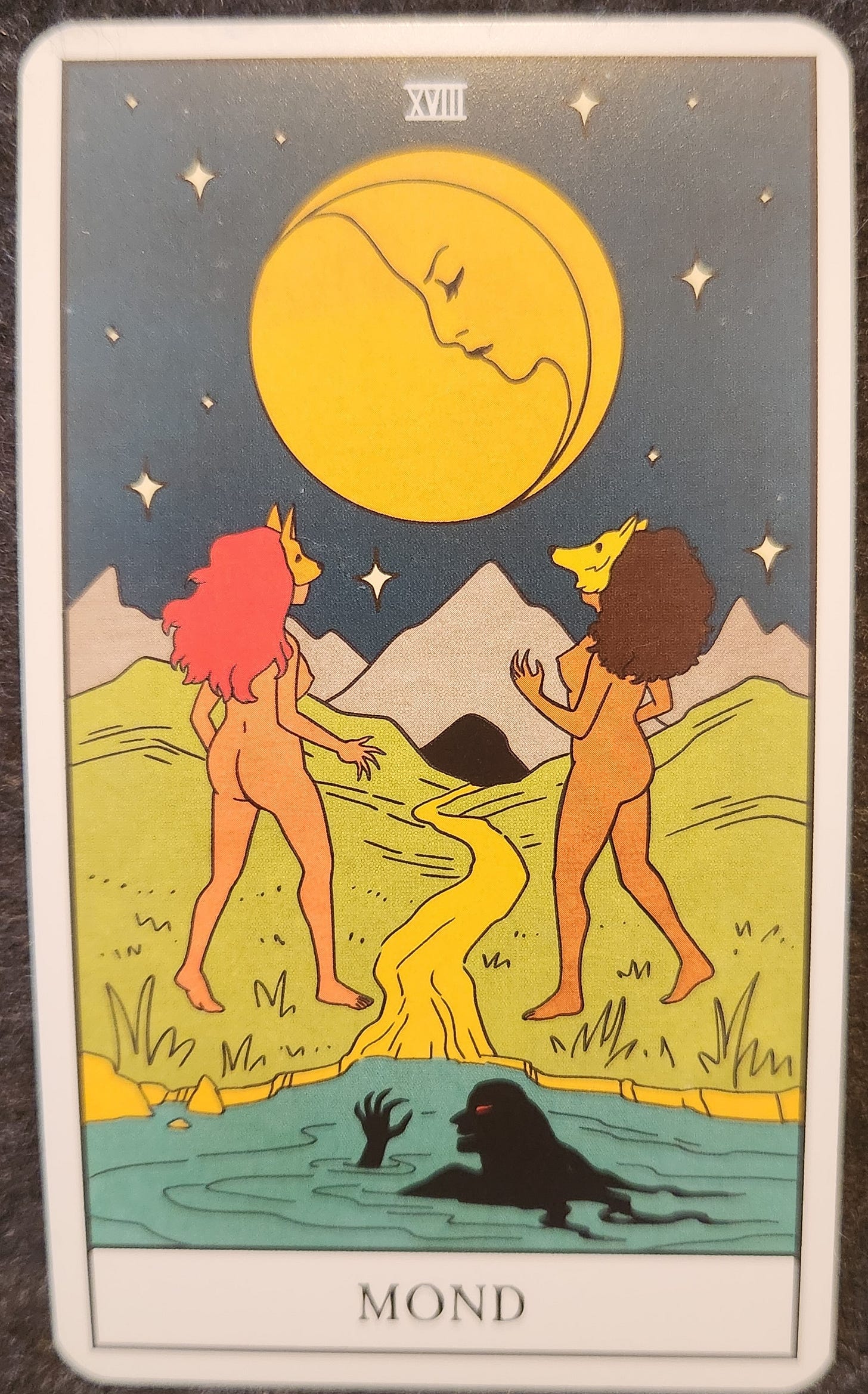 A tarot card labelled XVIII Mond. Two naked women in masks are looking up at a yellow moon hanging in the sky above. One of the women is wearing a dog mask. The other is wearing a wolf mask. Between the women, is a yellow stream of water. Behind them, is a lake with a shadowy female figure with a glowing red eye. 