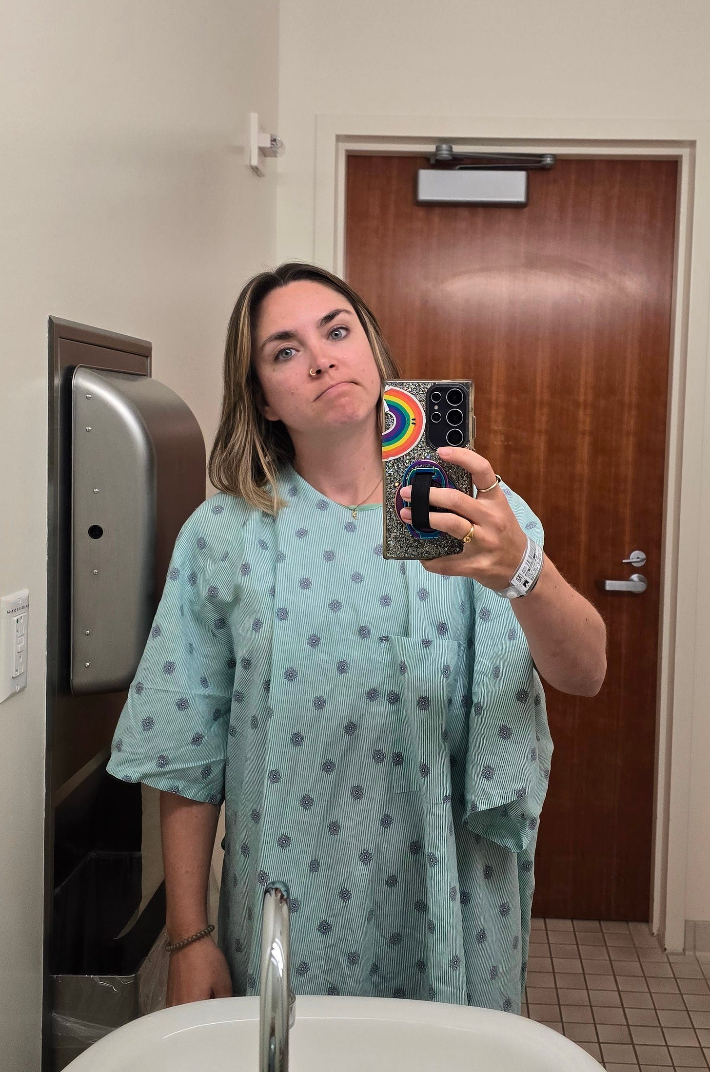 Cassidy in a hospital bathroom, wearing a hospital gown, frowning with the face of someone being so over medical appointments.