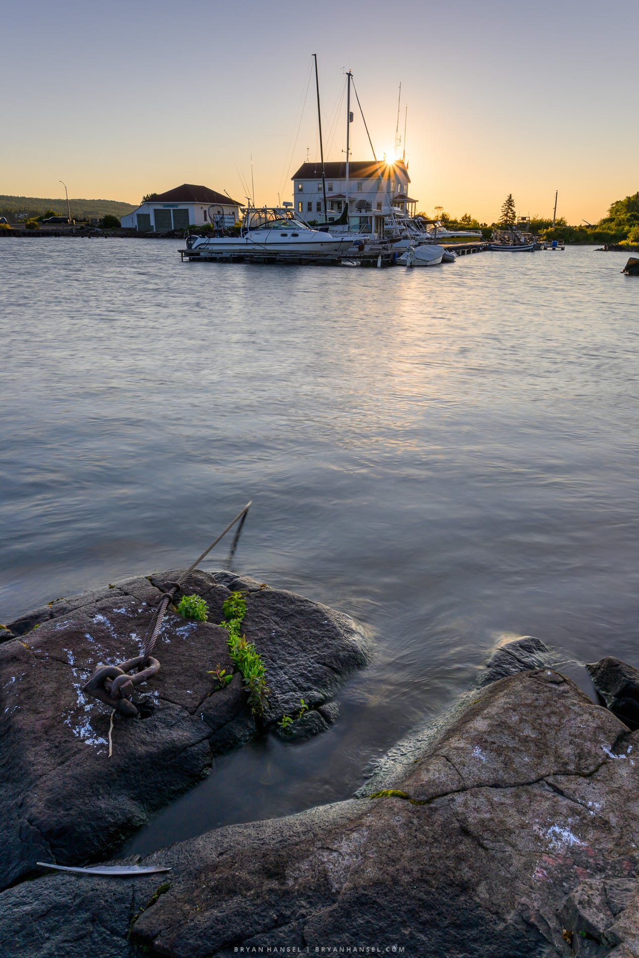Sunrise over the Grand Marais harbor, with boats moored in front of the Coast Guard building. Smooth water reflects the warm morning light, while granite shoreline rocks and a mooring chain fill the foreground.
