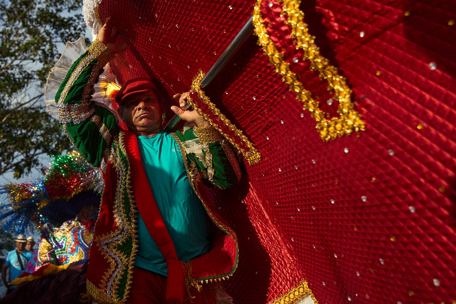 A person in festive, colorful clothing holds up part of a large, ornate red costume during a lively outdoor celebration or parade, with others in bright outfits in the background.