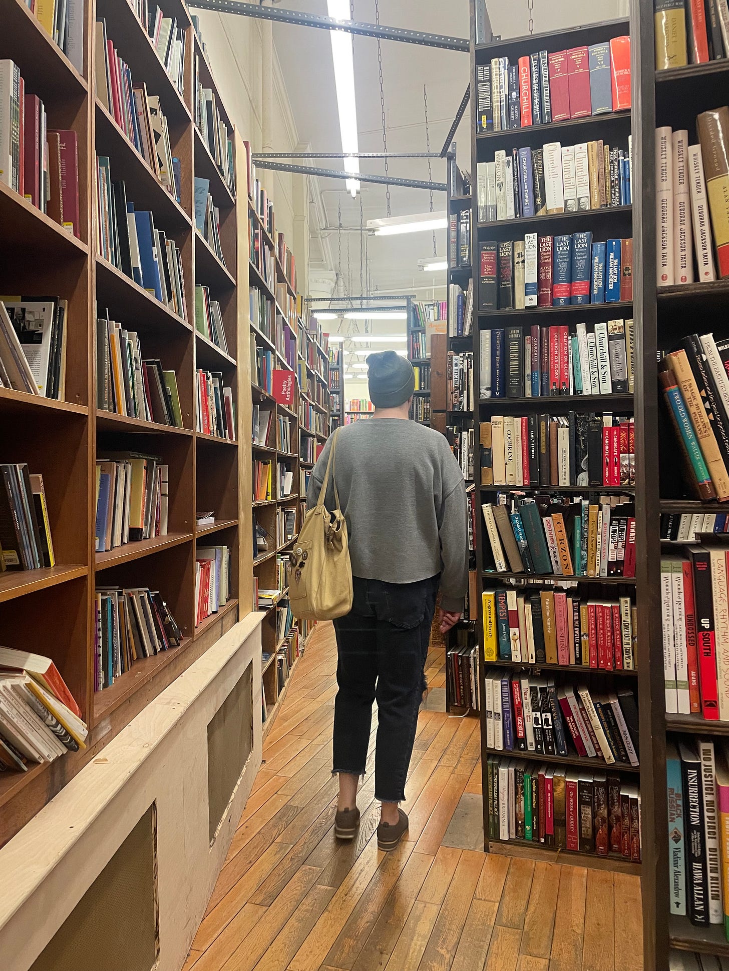 photo taken behind a person in black jeans, a gray sweatshirt, and a purple knit cap walking through stacks of books in a bookstore