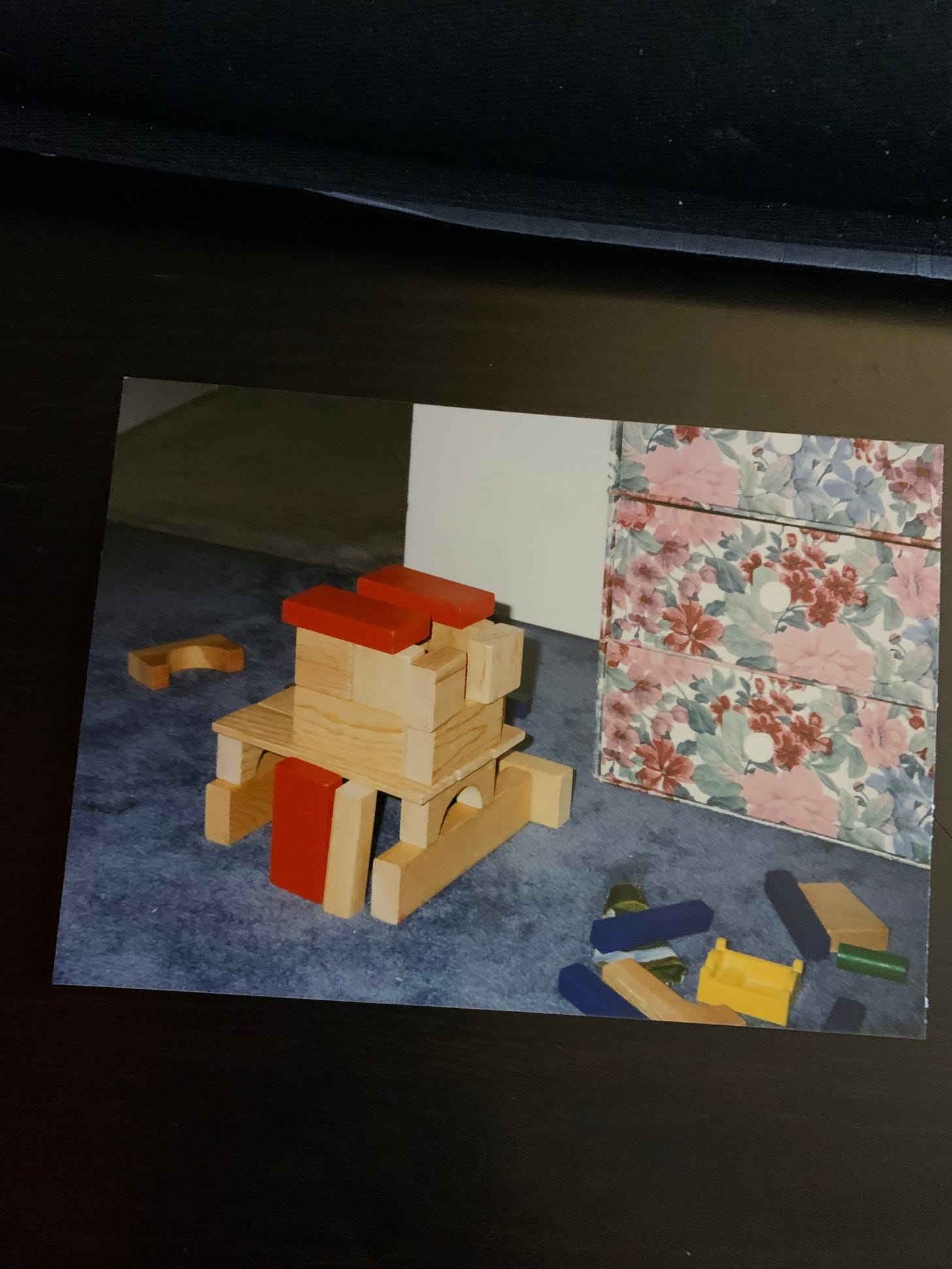wooden block 2 story house on top of a blue rug, in front of a small dresser with flower pattern