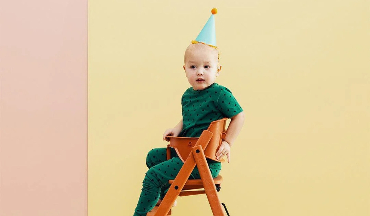 A child in a green polka-dot outfit sits in a wooden high chair, wearing a light blue party hat against a colourful backdrop.