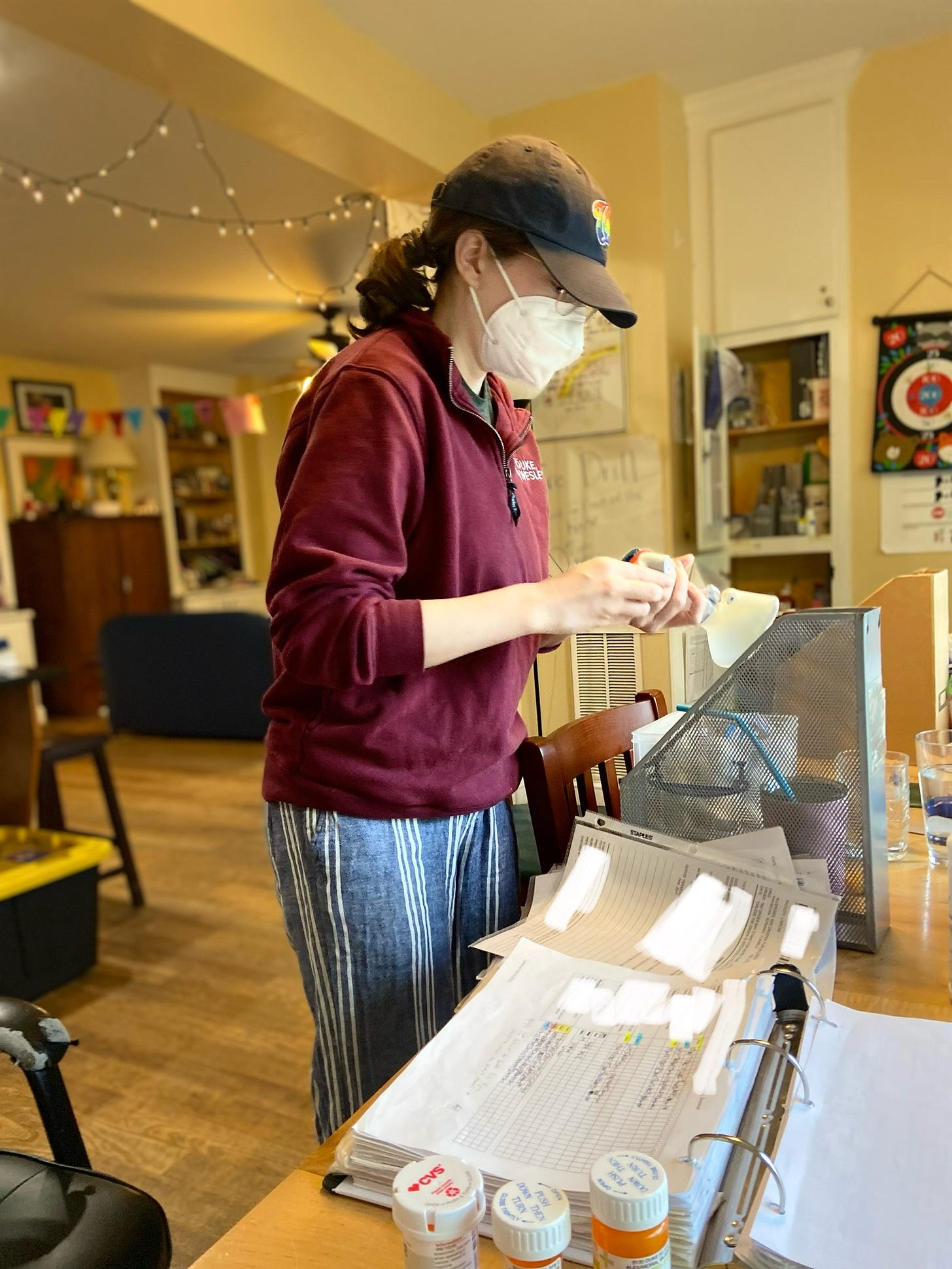 The author is wearing casual clothes and an N95 mask. She is checking medications at a table that includes a binder full of papers, an iced coffee, and more