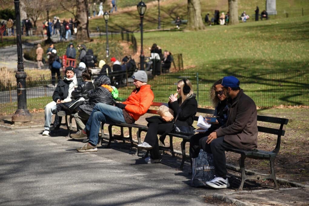 View of park bench with many people talking, reading, eating, bundled up against cold weather. Grass and other benches in view. 