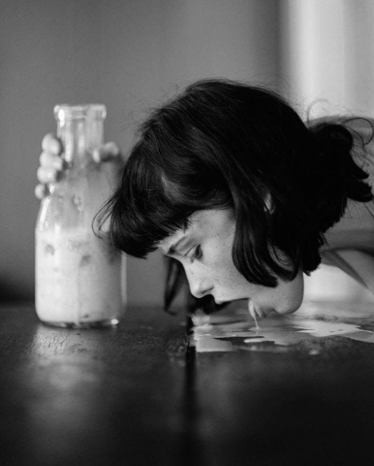 Black-and-white photo of a young woman with dark hair leaning down to lick spilled milk off a table. She holds a nearly empty glass bottle in one hand. The image is intimate, strange, and slightly unsettling.