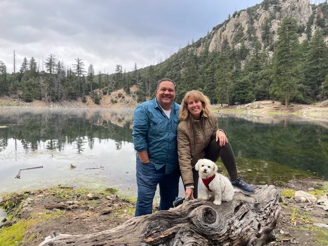 Mickey is wearing a teal shirt, Julie is wearing a brown jacket, and their little white dog Ellie is wearing a red harness, Ellie is sitting on a log by a small lake, Mickey and Julie are behind her, with pine trees and an overcast sky on the other side