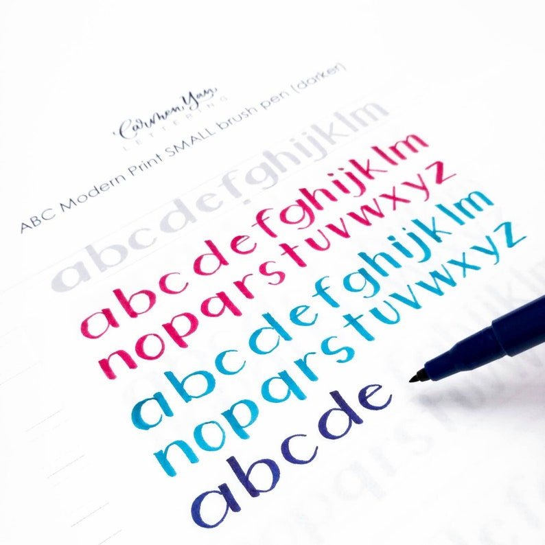 May include: A close-up of a white sheet of paper with the alphabet written in four different colors: gray, pink, blue, and dark blue. The letters are written in a modern print style. A blue brush pen is lying on the paper, with the tip of the pen resting on the last letter of the alphabet.