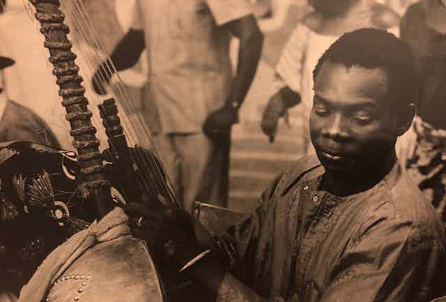 A sepia-toned vintage photograph of a young African man smiling to himself, eyes closed, as he plays a kora and figures are visible dancing behind him.