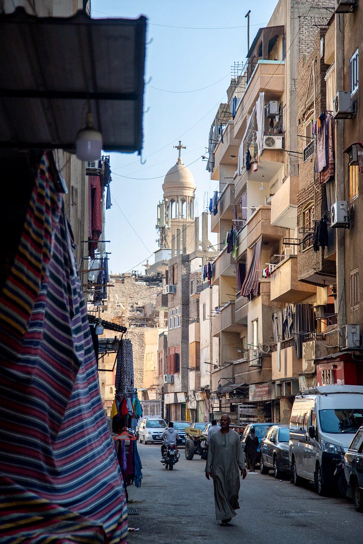 Top left: A narrow street in Luxor with apartment buildings, parked cars, and hanging textiles, with a Coptic church dome visible above the rooftops Top right: A woman in a black abaya buying fruit from a donkey-drawn cart loaded with oranges and bananas, parked in front of shuttered shopfronts Bottom right: A young man sitting inside a small vegetable stall framed by a green wall covered in Arabic calligraphy and religious posters, with piles of tomatoes, eggplant, and potatoes in the foreground Bottom left: A butcher in a blue plaid shirt bending over a cow's head on a cutting board, with a leg hanging from a hook and a Champions football poster on the red door behind him