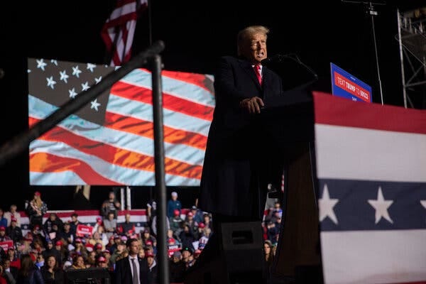 Former President Donald J. Trump speaking at a podium with an audience and a projection behind him.