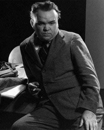 A black and white photo of a man in a suit sitting at a desk. He has a cigarette in his right hand and looks down to the right.