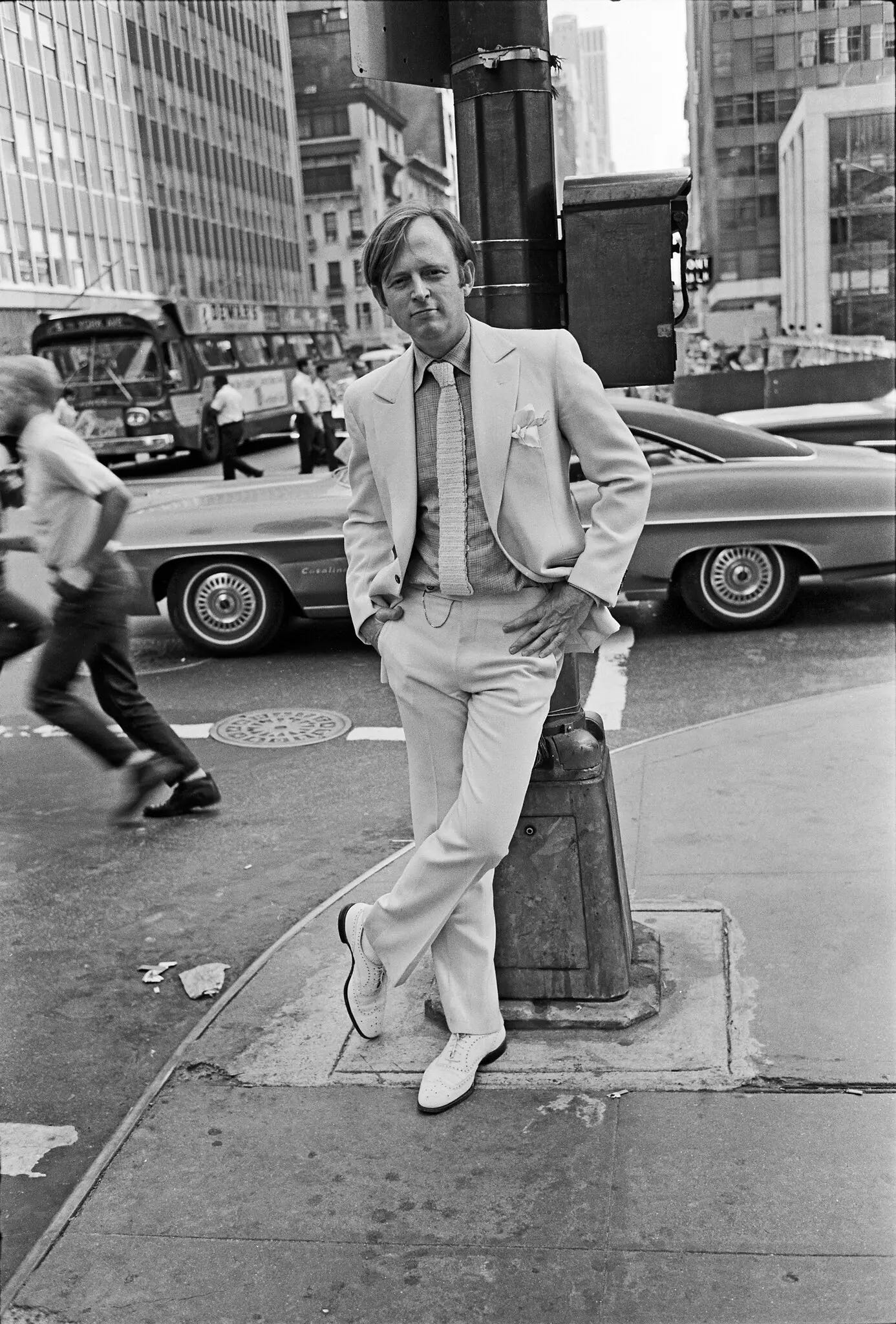 This black-and-white photo shows a clean-shaven young man in a light-colored suit and tie and white saddle shoes, posing nonchalantly against a streetlight at a busy crossroads in Midtown Manhattan. 