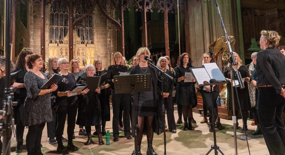 A group of choir singers holding books and standing on a stage, with a woman in front with a microphone