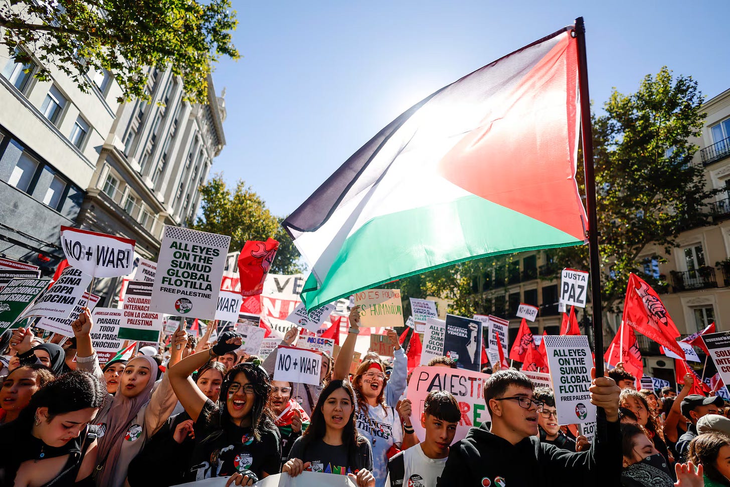 Una fotografia diurna scattata dal basso di una manifestazione in una strada cittadina assolata. In primo piano, una grande bandiera palestinese (nera, bianca, verde e rossa) è tenuta alta, quasi coprendo il sole e creando un forte bagliore sulla stoffa. La folla è composta da molte persone che sollevano cartelli e agitano le mani. Molti cartelli sono visibili con scritte in varie lingue, tra cui: "NO + WAR!", "ALL EYES ON THE SUMUD GLOBAL FLOTILLA FREE PALESTINE", "PAIEST RE", e "JUSTA PAZ" (Giusta Pace). Le persone in primo piano sorridono, gridano e alzano i pugni in segno di protesta. 