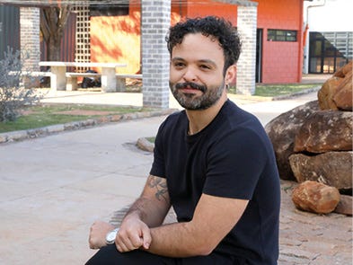 A imagem mostra um homem jovem de cabelos escuros e cacheados, com barba e bigode bem aparados, vestindo uma camiseta preta de mangas curtas. Ele está sentado em um ambiente externo, olhando para o lado com um sorriso discreto, revelando uma tatuagem no braço direito e um relógio de pulso. Ao fundo, há um pátio pavimentado, algumas pilastras de tijolos cinzas, uma parede laranja ao fundo e uma pilha de pedras grandes à direita. A imagem mostra um homem jovem de cabelos escuros e cacheados, com barba e bigode bem aparados, vestindo uma camiseta preta de mangas curtas. Ele está sentado em um ambiente externo, olhando para o lado com um sorriso discreto, revelando uma tatuagem no braço direito e um relógio de pulso. Ao fundo, há um pátio pavimentado, algumas pilastras de tijolos cinzas, uma parede laranja ao fundo e uma pilha de pedras grandes à direita.