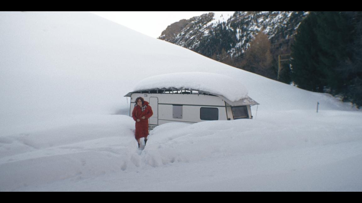 A bare-legged woman in a red coat walking away from a caravan in a snowedi-in valley