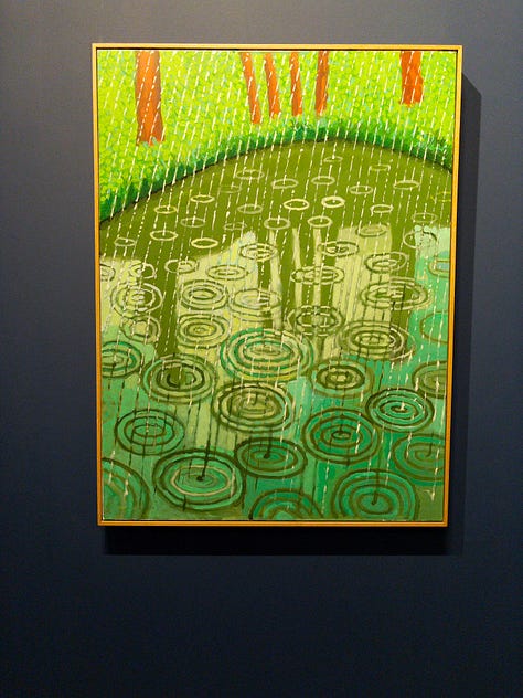 Multiple bright-colored paintings including a self-portrait of Hockney in a yellow plaid suit and big yellow glasses, the crowd viewing paintings, a mother and child in front of a bright painting of a massive tree, an annunciation painting redone in neon colors, a green lawn with sprinkler spray, a photograph of hockney working at his drawing table with his little terrier dog under the table and two paintings with raindrops hitting puddles among trees