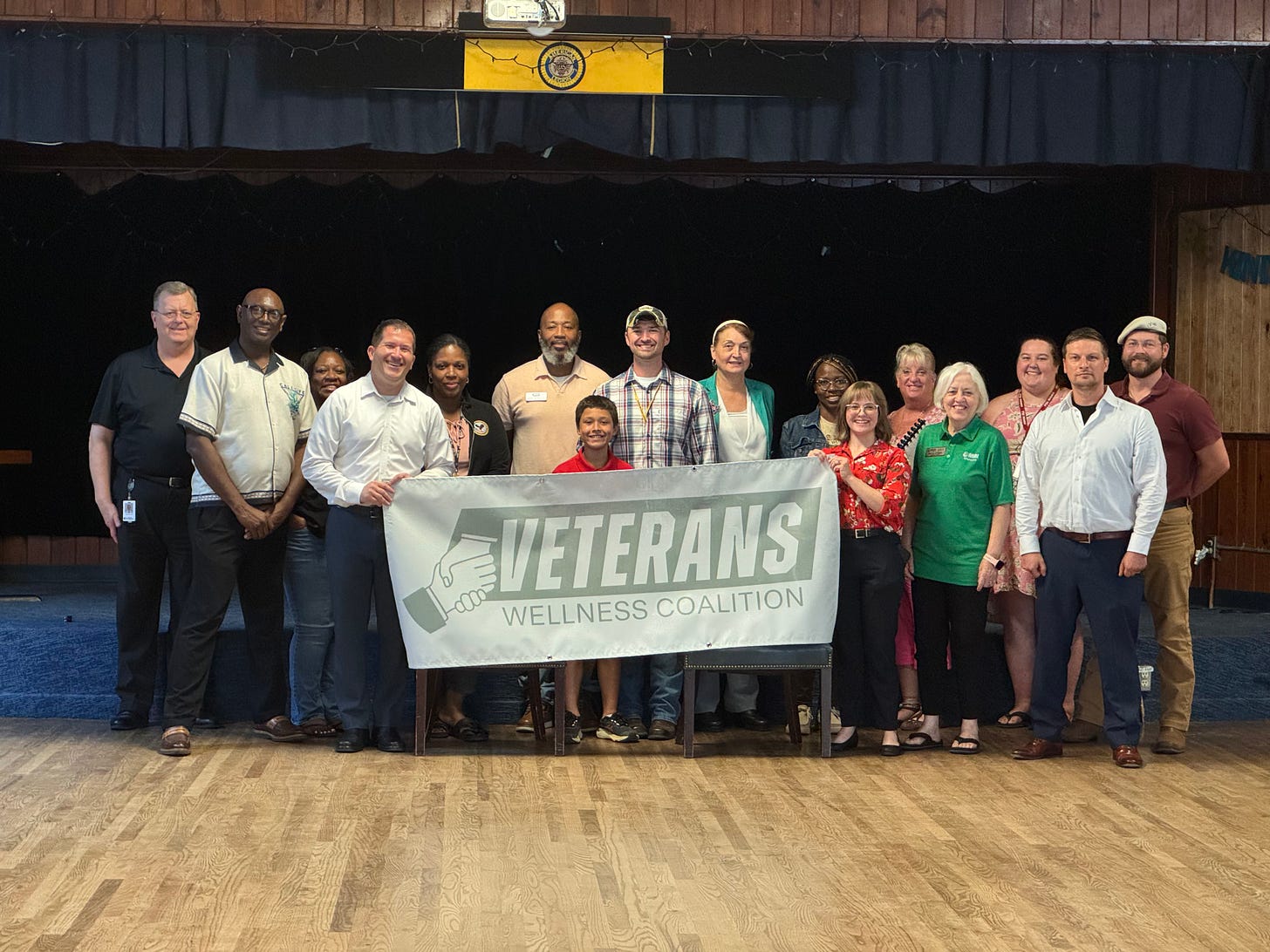 A group of people are standing together indoors on a wooden floor, posing for a group photo. They are holding a large sign that reads "VETERANS WELLNESS COALITION" with a handshake logo. There are about sixteen adults and one child in the group. In the background, a stage with dark curtains and a banner with a seal is visible. The people are smiling and appear to be part of an organized event supporting veterans' wellness. No specific recognizable figures, landmarks, or brands aside from the coalition’s name are visible. A group of people are standing together indoors on a wooden floor, posing for a group photo. They are holding a large sign that reads "VETERANS WELLNESS COALITION" with a handshake logo. There are about sixteen adults and one child in the group. In the background, a stage with dark curtains and a banner with a seal is visible. The people are smiling and appear to be part of an organized event supporting veterans' wellness. No specific recognizable figures, landmarks, or brands aside from the coalition’s name are visible.