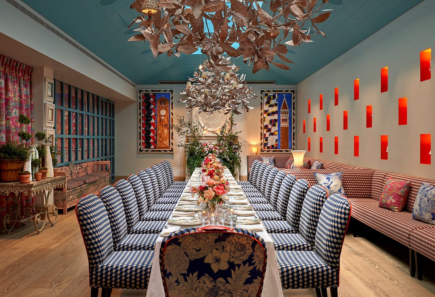 The Orangery at Warren Street Hotel with a vaulted blue ceiling and skylights. Banquette seating runs down the right-hand side of the room and a long table runs down the centre of the room with chairs upholstered in a blue, white and red patterned fabric. Two statement chandeliers hang from the ceiling and at the end of the room is a fireplace filled with foliage.