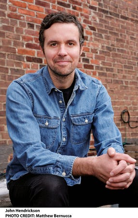 The author, in a denim button-up shirt and black pants, sits in front of a brick wall. The author, in a denim button-up shirt and black pants, sits in front of a brick wall.
