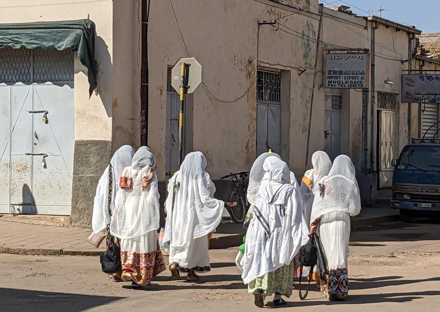Seven women are walking on a dusty city street. We only see their backs and their stark white shema (shawls) with colorful embroidery on the edges. Seven women are walking on a dusty city street. We only see their backs and their stark white shema (shawls) with colorful embroidery on the edges.