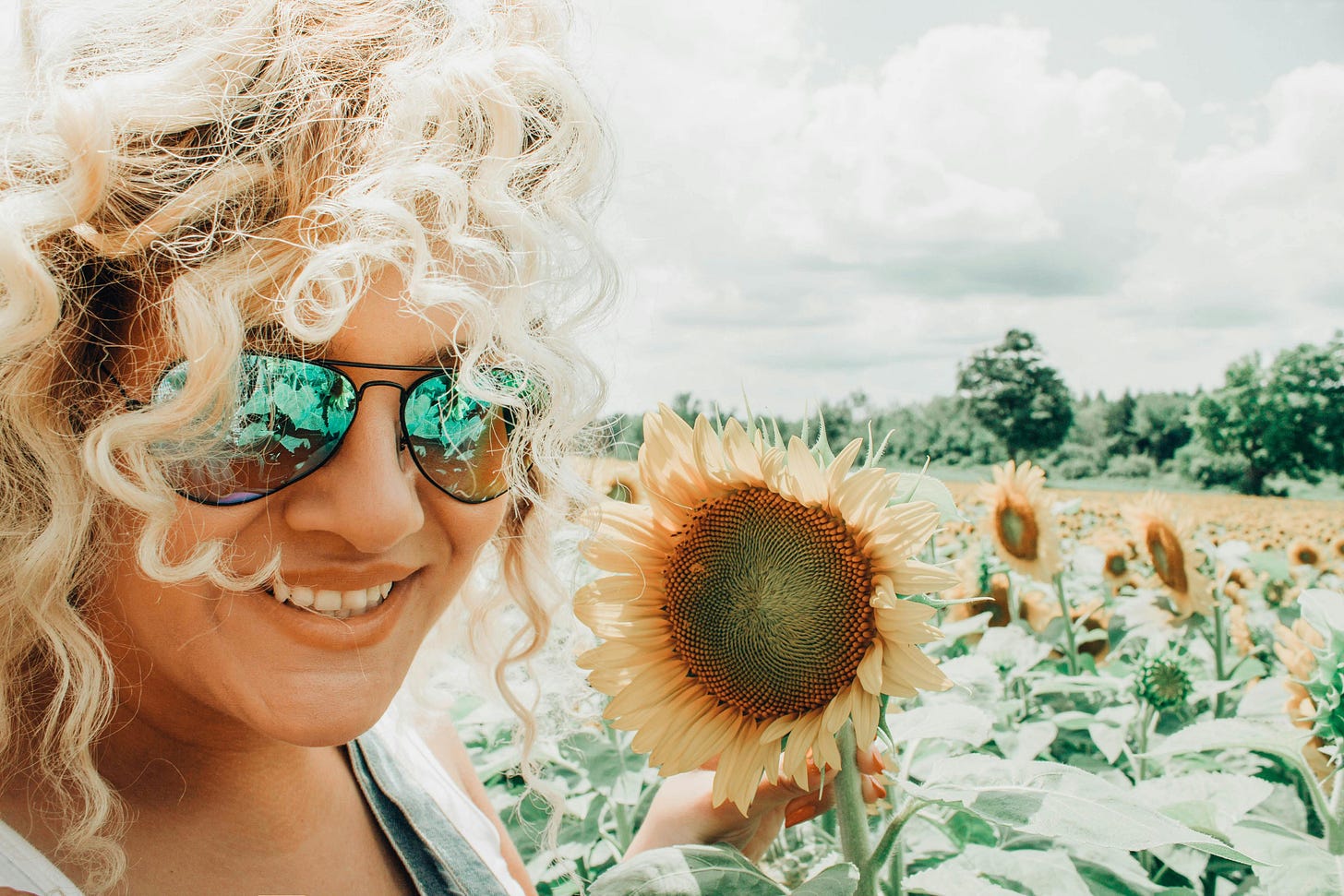 A woman with blonde hair and sunglasses stands in a field of sunflowers, hold the head of one in her hand and smiling at the camera. A woman with blonde hair and sunglasses stands in a field of sunflowers, hold the head of one in her hand and smiling at the camera.