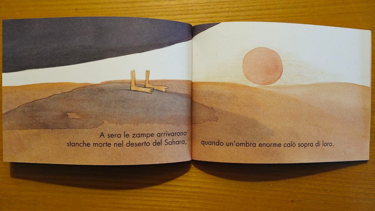 Due tavole dell'albo illustrato "Urla di mamma" appoggiato su un tavolo di legno: nelle due pagine l'illustrazione di un deserto con due zampe di pinguino sovrastate da qualcosa di nero; compare la scritta 'A sera le zampe arrivarono stanche morte nel deserto del Sahara, quando un'ombra enorme calò sopra di loro.'