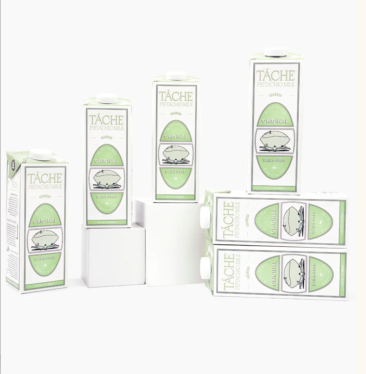 six cartons of Tache pistachio milk, artfully posed like they're high fashion models, some casually lying on their side, others on pedestals