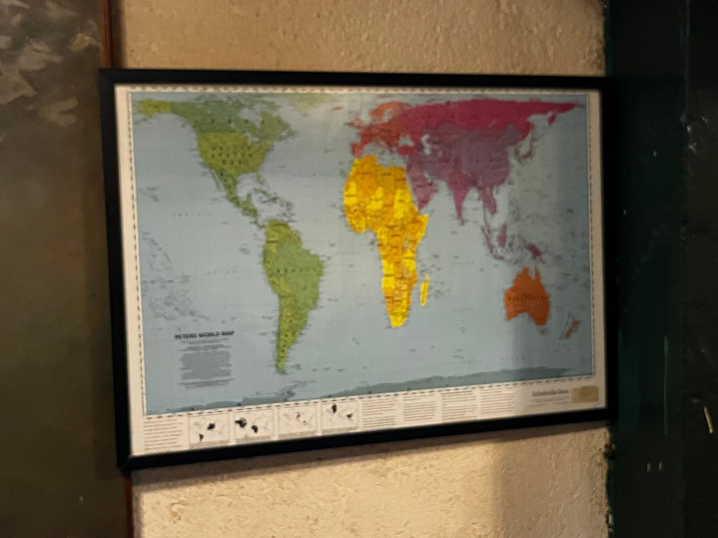 A framed world map hangs on a textured beige wall in a dimly lit interior. The map uses a Peters projection, with continents color-coded: the Americas in green, Africa in yellow and visually dominant at center, Europe and Asia in red and purple tones, Australia in orange. The lower left corner of the map reads "Peters World Map." A dark doorframe is visible at the right edge of the image.