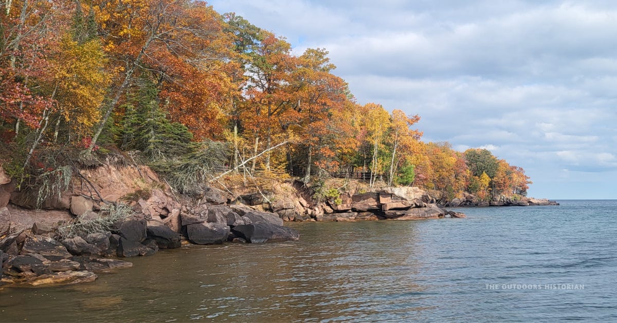 Rocky red sandstone shoreline along Lake Superior lined with trees in peak fall color — orange, red, and gold leaves against a partly cloudy sky.