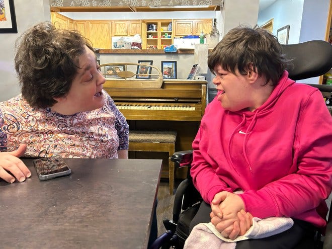 A woman and a young adult with disabilities are engaged in a lively conversation at a table, with a piano visible in the background.